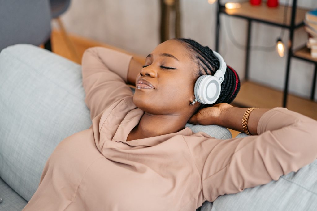 Young beautiful black woman listening to guided meditation on headphones while relaxing in the living room at home.